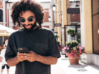 Handsome smiling hipster  model. Sexy unshaven Arabian man dressed in summer clothes. Fashion male with long curly hairstyle using smartphone apps, looking at cellphone screen. Holds phone
