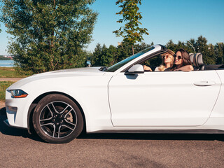 Portrait of two young beautiful and smiling hipster female in convertible car. Sexy carefree women driving cabriolet. Positive models riding and having fun in sunglasses outdoors. Enjoying summer days