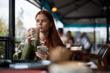 Woman sitting in a cafe drinking coffee from a mug with phone in hand, blogger photographing food content for social media, portrait of a stylish girl with red hair in the autumn