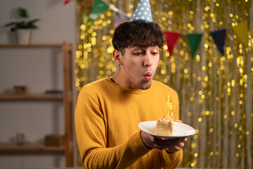 Excited man celebrating birthday at home, blowing candle on cake, wearing party hat and having fun, standing over golden foil background. Holidays and celebration