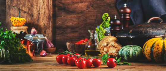 Cooking food background with tomatoes, pumpkins, celery, herbs on rustic wooden kitchen table with cast iron pan, spice grinders, cutting board, spices and seasonings, storage jars. Banner
