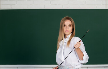 Serious teacher pointing on lesson. Cute young woman with pointer teaching near blackboard.