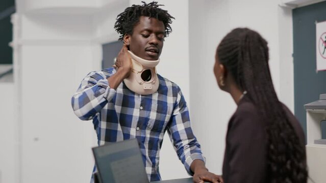 Patient with neck collar at reception counter talking to medical worker, asking for assistance. Injured man with cervical brace waiting to attend checkup appointment with spcialist. Handheld shot.