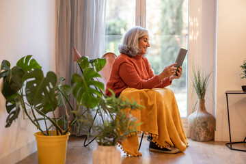 senior woman with gray hair, in the living room at home sitting in an armchair by the window, the...