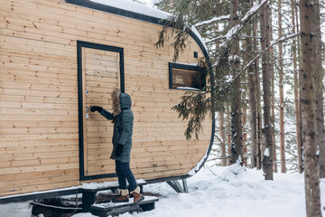 Girl walking in winter snowy forest near wooden tiny house. Winter landscape. Glamping