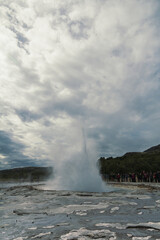 Tourists enjoy geyser eruption landscape photo. Beautiful nature scenery photography with grey sky on background. Idyllic scene. High quality picture for wallpaper, travel blog, magazine, article