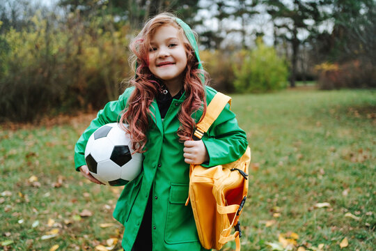 Fototapeta Smiling girl standing with backpack and soccer ball at park