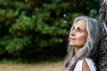 Thoughtful woman with gray hair leaning on tree trunk at forest