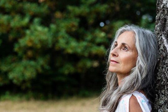 Thoughtful woman with gray hair leaning on tree trunk at forest