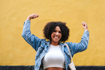 Cheerful young woman flexing muscles in front of wall