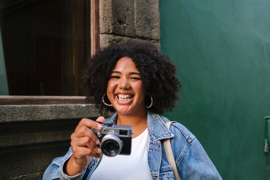Cheerful young woman with curly hair holding analog camera in front of wall