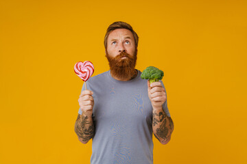 Bearded man holding lollipop and broccoli standing over background