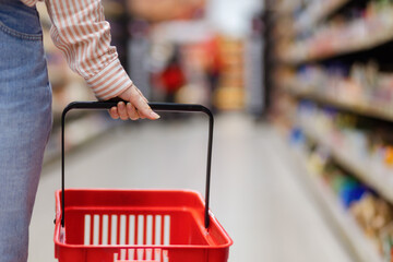 Female hand holds empty red grocery cart, close-up. In background is shopping mall with shelves in...