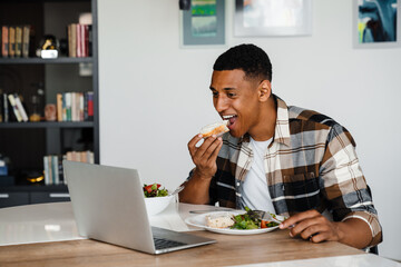 Young african man watching movie on laptop while having lunch at home
