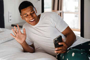 Positive african man waving while having video call via mobile phone lying in bed