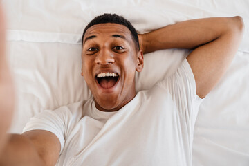 Top view of joyful screaming african man taking selfie while lying in bed