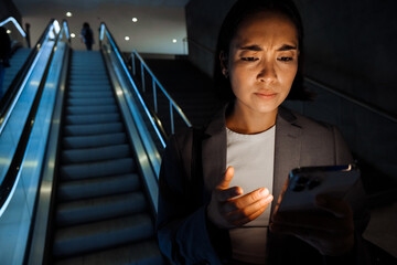 Young asian woman frowning while using smartphone standing on escalator indoors
