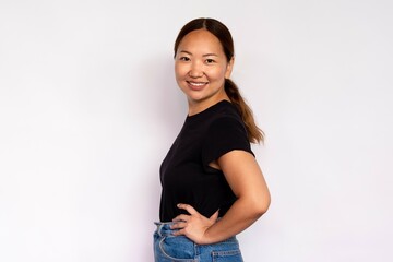 Portrait of attractive young woman smiling at camera over white background. Asian lady wearing black T-shirt and jeans posing with hands on hips. Female beauty concept