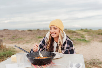 Lifestyle image of happy traveler woman eating on a table outdoor along beach. Nomad and travel lifestyle concept