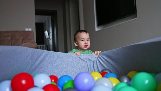 Active Caucasian baby boy playing near the dry pool filled with balls. Kid jumps up holding by the basin edge.