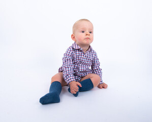 Playful beautiful baby boy wearing plaid bodysuit standing over white studio background sitting and smiling to the camera. Baby boy wants to play with his mother 