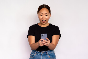 Portrait of amazed young woman reading news on mobile phone over white background. Asian lady wearing black T-shirt and jeans reading message on smartphone. Mobile communication concept