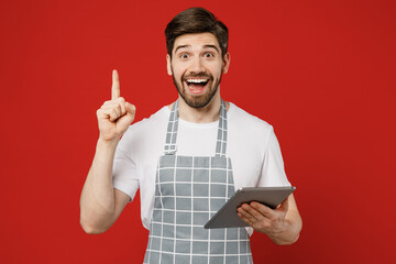 Young fun male housewife housekeeper chef cook baker man wear grey apron hold use digital tablet pc computer point index finger up isolated on plain red color background studio. Cooking food concept.