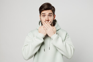 Young scared sad surprised shocked unshaven caucasian man wears mint hoody looking camera covering mouth with hand isolated on plain solid white background studio portrait. People lifestyle concept.