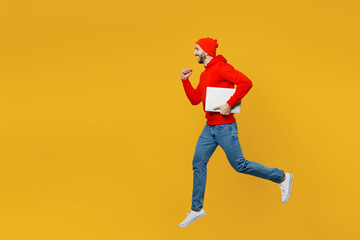 Fototapeta na wymiar Full body side profile view young man wear red hoody hat jump high hold closed laptop pc computer run fast hurry up isolated on plain yellow color background studio portrait. People lifestyle concept.