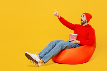 Full body young surprised shocked man in red hoody hat sit in bag chair hold inhand takeaway popcorn bucket watch movie film point index finger aside on area isolated on plain yellow color background.