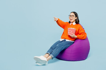 Full body fun young woman of Asian ethnicity in orange sweater glasses sit in bag chair hold...