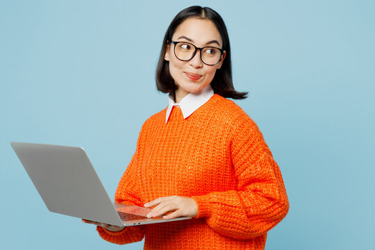 Young smart IT woman of Asian ethnicity wear orange sweater glasses hold use work on laptop pc computer look aside on area isolated on plain pastel light blue cyan background People lifestyle concept