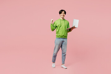 Full body young IT man of Asian ethnicity in green hoody hold use work on laptop pc computer do winner gesture isolated on plain pastel light pink background studio portrait People lifestyle concept