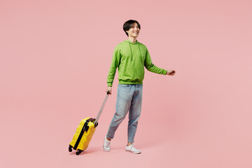 Full body traveler man of Asian ethnicity wear green hoody hold suitcase valise isolated on plain pastel pink background Tourist travel abroad in free spare time rest getaway. Air flight trip concept