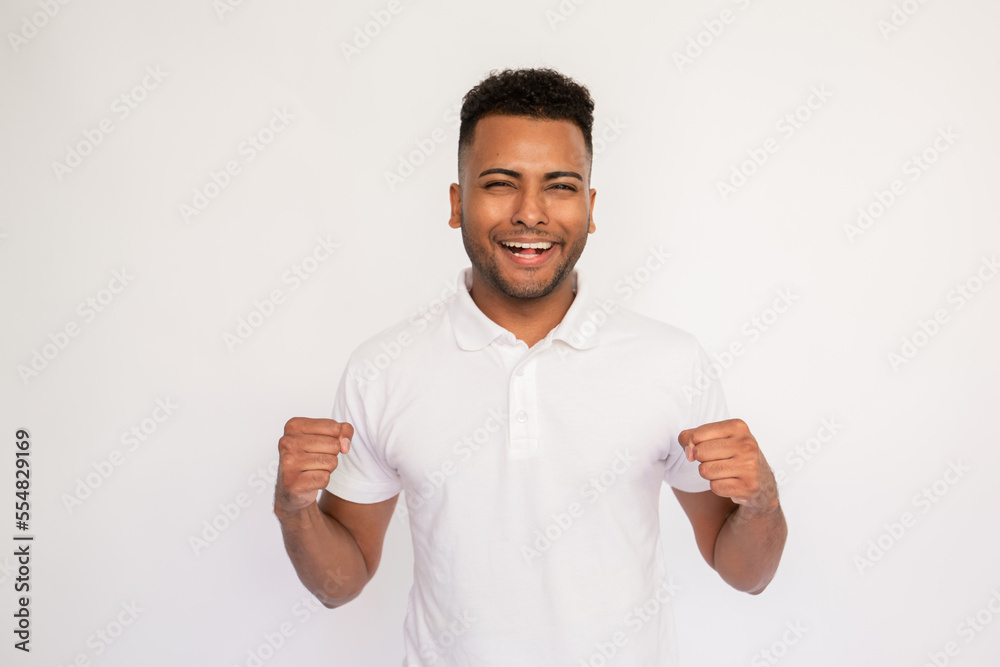 Wall mural Joyful young man celebrating win. Male Indian model with brown eyes and curly hair in white polo shirt enjoying successful result. Winner, achievement concept