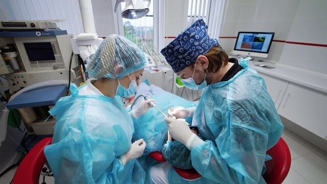 Patient wearing cap, dark glasses and covered with sheet lies on the chair. Dentist performing tooth treatment assisted by a nurse.