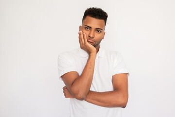 Bored young man holding his face with hand. Male Indian model with brown eyes and curly hair in white polo shirt looking bored hearing something uninteresting. Boredom, dissatisfaction concept