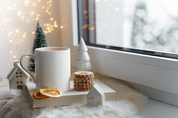 Eine weiße Tasse Tee und Kekse auf einem Buch an einem Fenster. Weihnachten, Lichterkette.