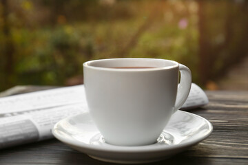 White cup with coffee and newspaper on wooden table, closeup. Morning ritual