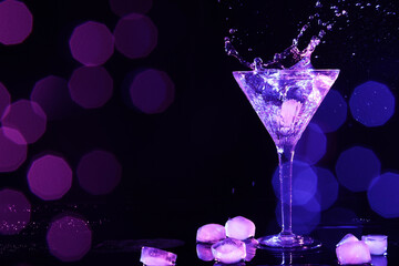 Glass of martini with ice and splashes in neon lights on dark background, bokeh effect. Space for text