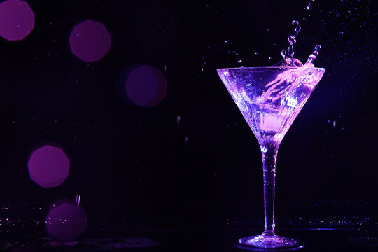 Glass of martini with ice and splashes in neon lights on dark background, bokeh effect. Space for text