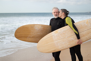 Happy elderly couple going in for surfing together. Sporty man and woman in wetsuits standing on...