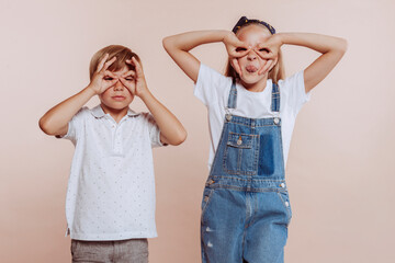 Young brother and sister doing ok gesture like binoculars with hands, sticking tongue out, eyes looking through fingers. funny expression. Studio portrait isolated over beige background. Children.