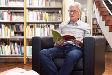 Portrait of serious old man sitting with book in library. Thoughtful man reading in arm-chair enjoying searching information to improve knowledges about computers. Education for adult people concept
