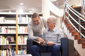 Portrait of happy men having fun while reading. Smiling aged man and his young teacher studying in library discussing books and tasks and joking. Modern education and studying for adult people concept