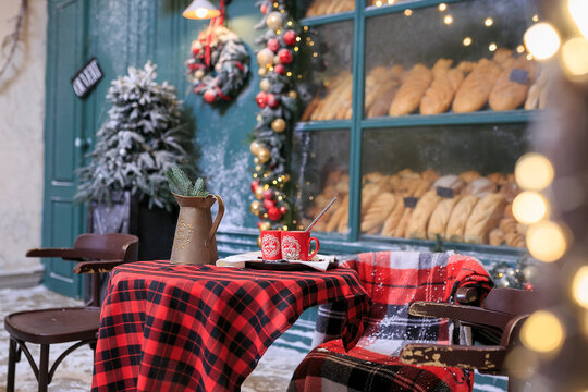 Table for two. Christmas bakery exterior with Christmas decorations and fresh bread in glass showcase. Cozy outdoor cafe on Christmas eve. Copy space. seasonal greetings concepts. Red cups of coffee.