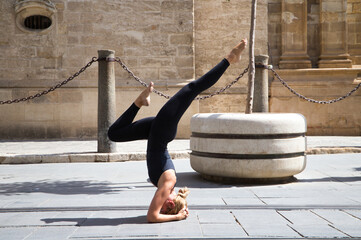 Middle-aged woman doing the yoga pose sirsasana, headstand. She is on the street between the...