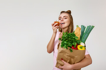 woman with paper bag with vegetables biting apple.