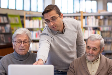 Portrait of positive elderly men with teacher in library. Young instructor teaching grey-haired men to use computer and to find information when studying. Education for adult people concept