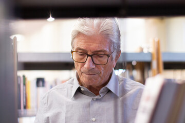 Portrait of serious elderly man. Thoughtful Caucasian man with grey hair standing near bookshelves choosing, reading books about computer technologies with interest. Education for adult people concept
